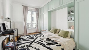 Quarto com cama, escrivaninha com luminária, janela com cortinas e varanda.