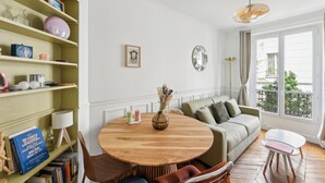 Sala de estar com uma mesa redonda de madeira, um sofá verde e uma poltrona branca. Há uma estante com diversos objetos e uma janela com vista para o verde.