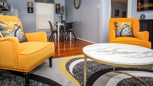 A living room with two yellow armchairs, a round coffee table, a dining table with chairs, a clock on the wall, and a TV mounted on the wall. - Downtown Cozy 2 BR Apt - 4min drive MardiGras Park (Mobile)
