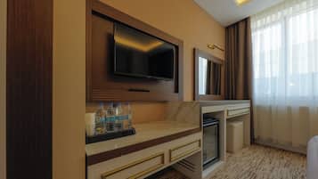 A hotel room with a flat-screen TV mounted on the wall, a minibar, a small refrigerator, and a bed with a white headboard.