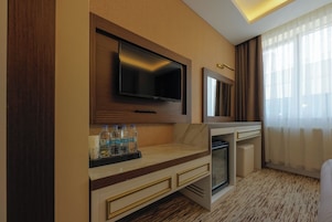 Habitación de hotel con televisión de pantalla plana montada en la pared, un minibar, un refrigerador pequeño y una cama con cabecera blanca.