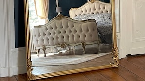A large ornate mirror with a gold frame reflecting a bedroom with a tufted headboard, a chandelier, and a patterned bedspread.