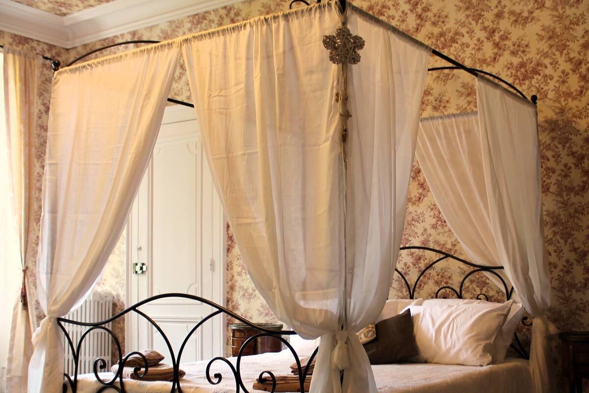 Un lit à baldaquin à quatre poteaux, avec une structure en métal, un couvre-lit blanc et un papier peint à motifs floraux.