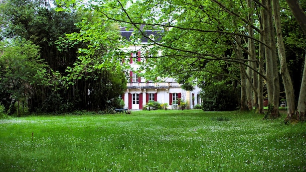 A large, white building with red shutters, surrounded by lush green trees and grass.
