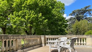 A stone balustrade with white wicker furniture on a paved area, surrounded by lush green trees.