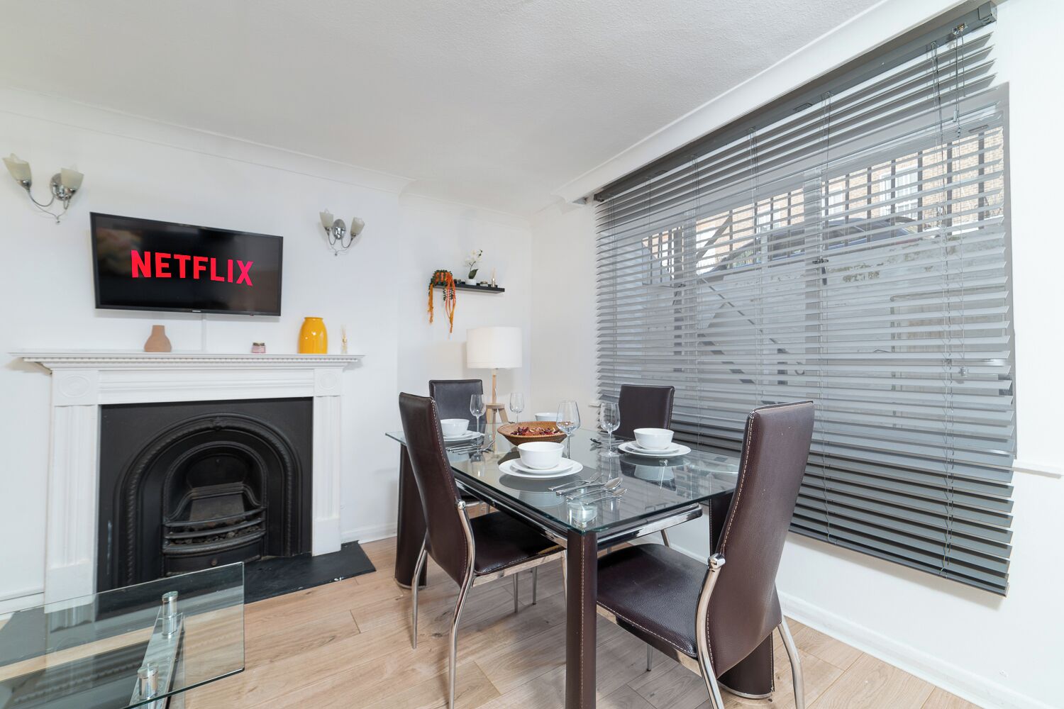 Un comedor moderno con una mesa de vidrio, sillas de cuero, chimenea y televisor empotrado mostrando 'NETFLIX'.