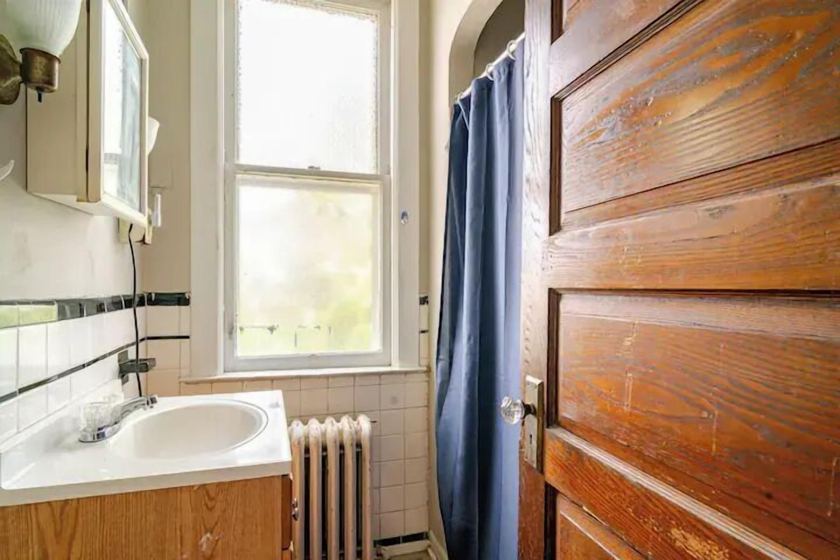Een badkamer met een houten deur, een wit wastafel, een raam en een radiator.