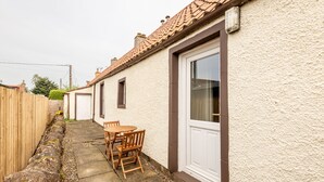 A residential property with a paved patio, wooden furniture, and a white door. - Jura Cottage | Peat Inn (Peat Inn)