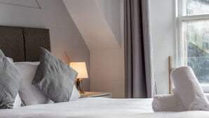A hotel room with a bed, pillows, a nightstand with a lamp, and a window with curtains. - Spire View Apartment (St Andrews)