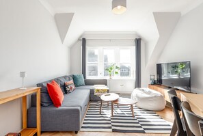 A modern living room with a grey sofa, a wooden coffee table, a bean bag, a TV, and a large window with curtains.