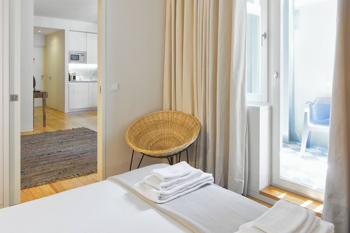 A hotel room with a bed, a wicker chair, a view of the outside, and a kitchen in the background.
