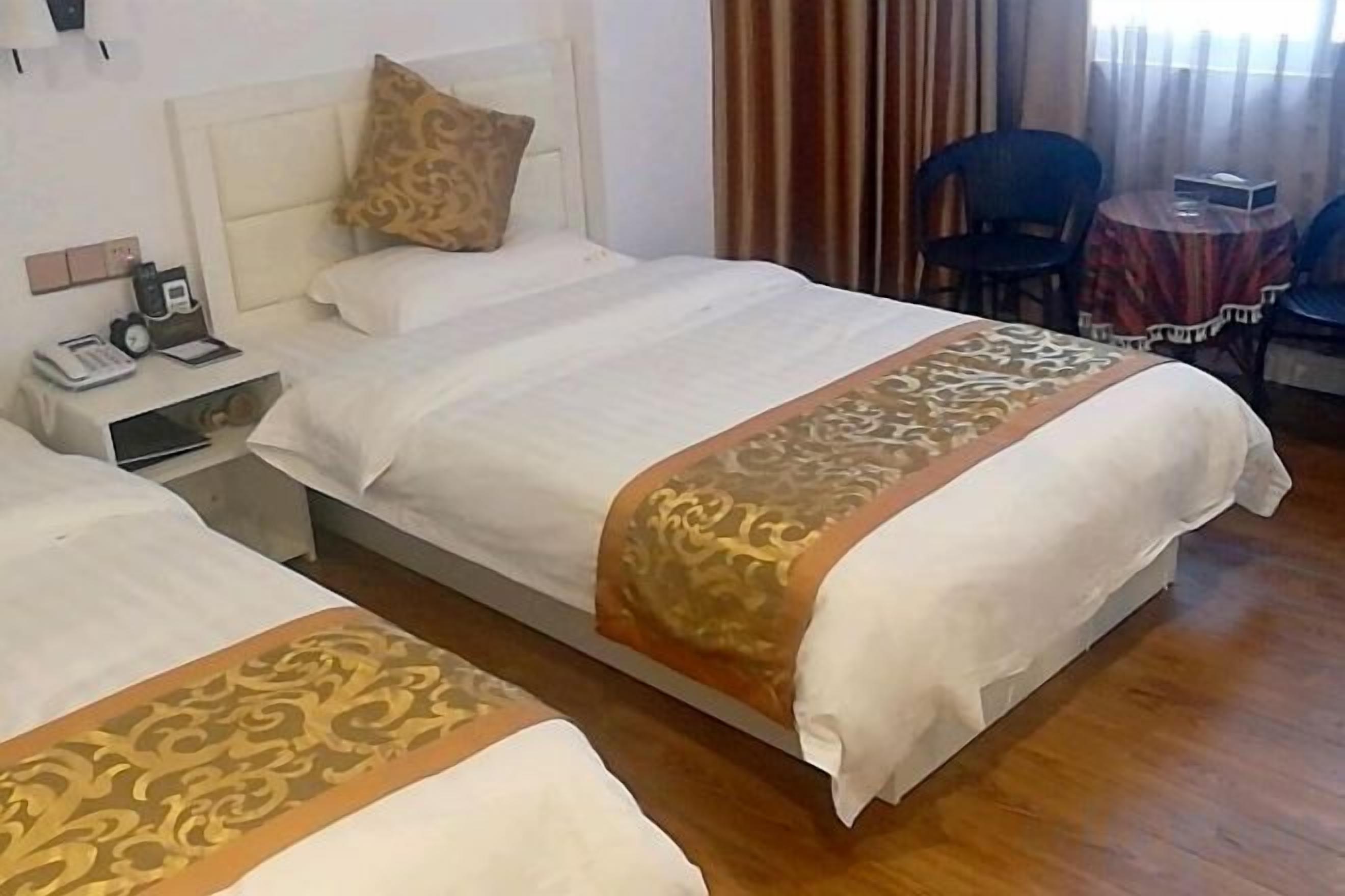 Dos camas individuales con sábanas blancas y cabeceros decorativos, una mesita de noche con teléfono y una pequeña mesa con una silla en la habitación.