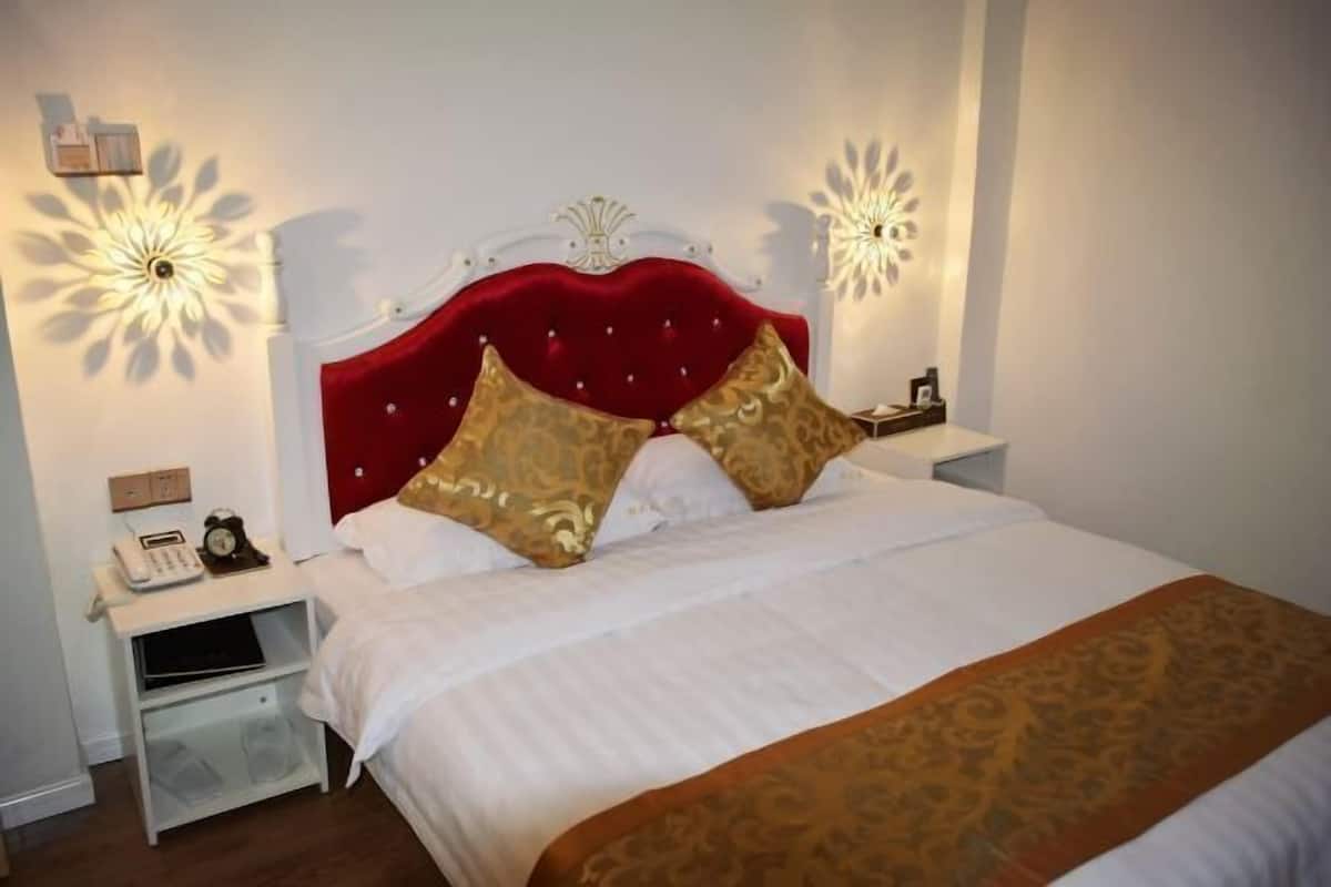 Una cama bien hecha con cabecera roja, ropa de cama blanca y una manta marrón. Hay dos mesitas de noche con lámparas, un reloj y una pequeña estantería.