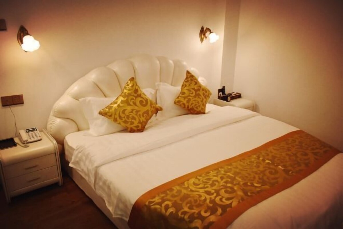 Habitación de hotel con una cama grande, ropa blanca y dos almohadas decorativas. Hay una mesita de noche con un teléfono y una lámpara.
