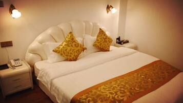 A hotel room with a large bed, white bedding, and two decorative pillows. There is a bedside table with a phone and a lamp.
