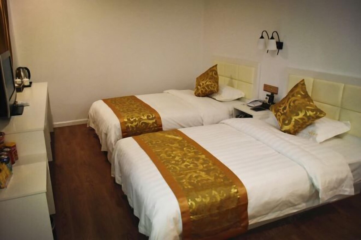 Habitación de hotel con dos camas, cada una con ropa de cama blanca y detalles dorados. Hay una mesita de noche con una lámpara, una pequeña mesa con un teléfono y una lámpara de pared.