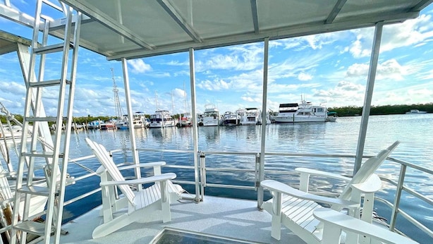 Une terrasse couverte avec des fauteuils Adirondack blancs, un plancher de verre et une vue sur les bateaux amarrés dans l’eau.