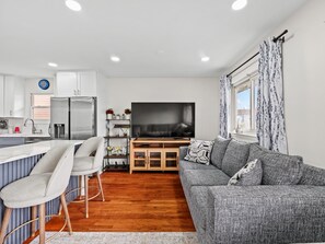 A modern kitchen with a built-in fridge, a dining area with bar stools, a grey sofa, and a TV on a wooden stand.