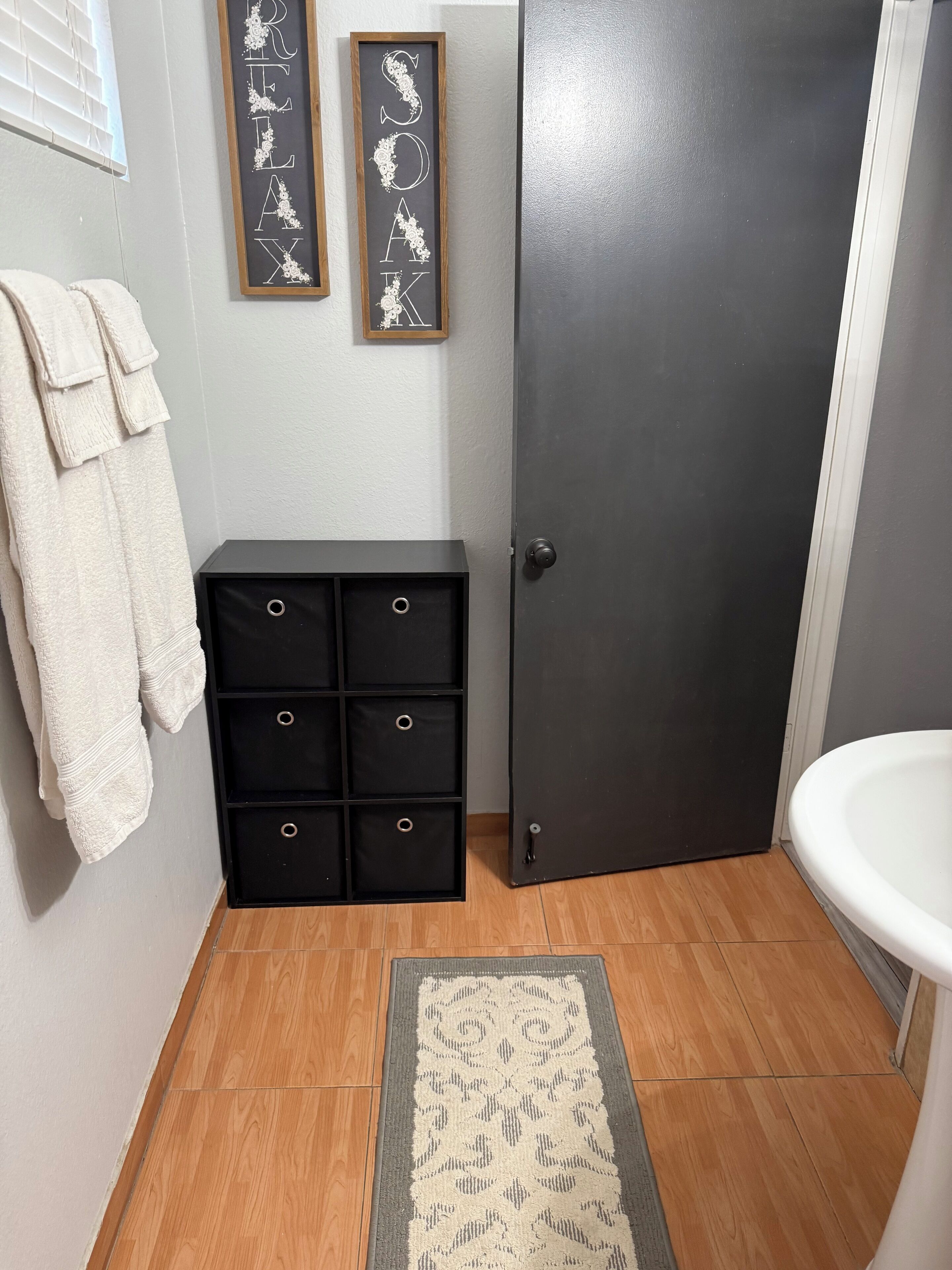 A bathroom with a wooden floor, a white sink, a dark door, and a black storage unit with drawers.