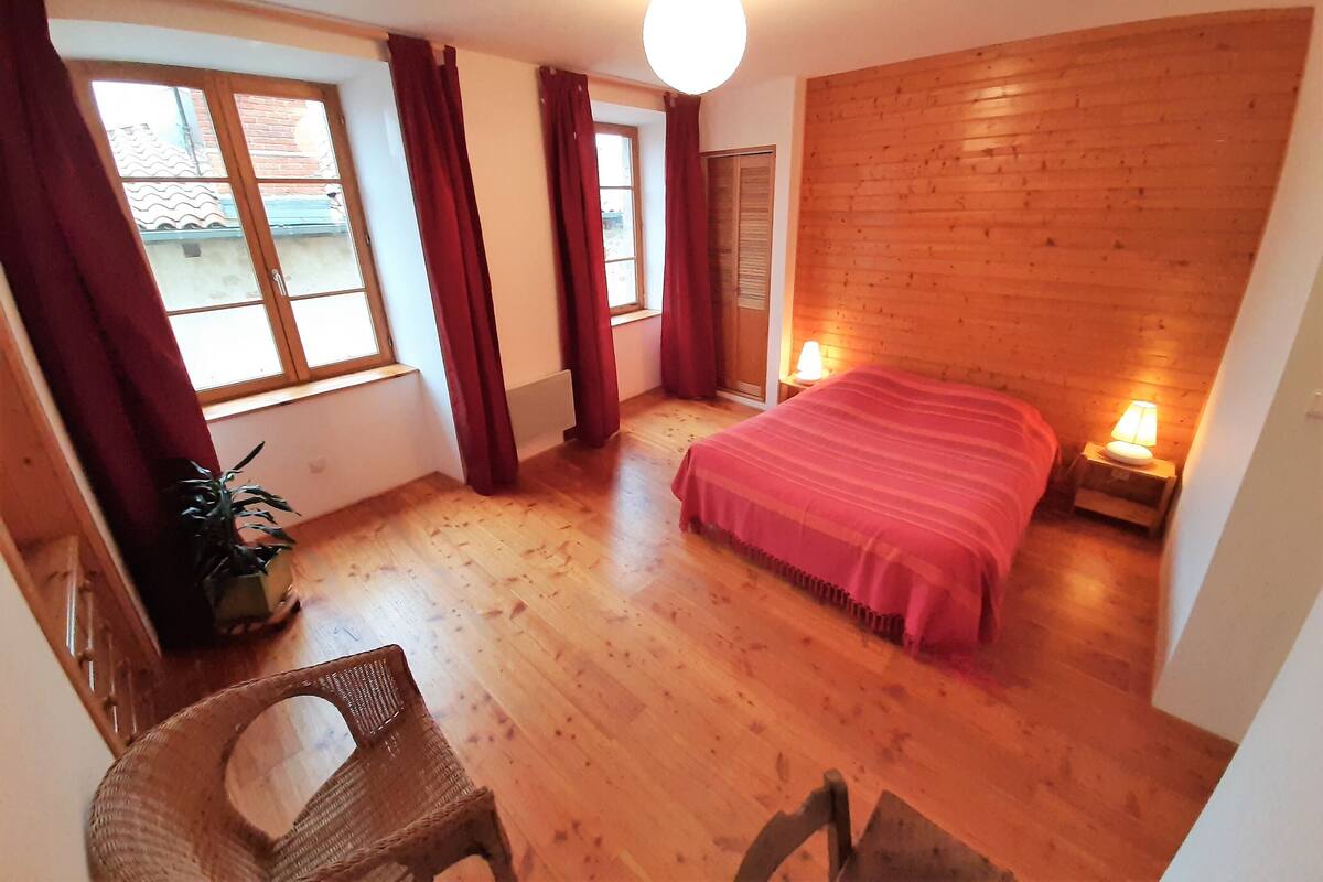 Een kamer met een houten vloer, een bed met een rode deken, twee bedlampjes, een raam met gordijnen en een stoel.