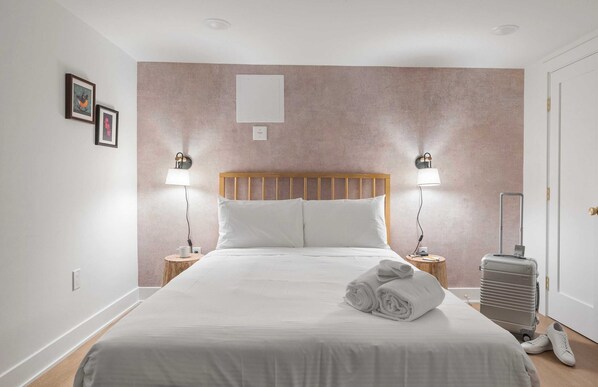 A neatly made bed with white linens, a wooden headboard, and two bedside lamps. A suitcase and a pair of shoes are on the floor beside the bed.
