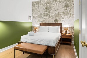 Ein ordentlich bezogenes Bett mit weißen Leinen, ein hölzerner Kopfteil und eine Sitzbank am Fußende. Zwei Nachttische mit Lampen, und eine Wand mit floralem Wandpapier.