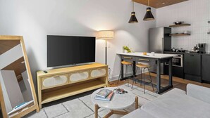 A modern living room with a flat-screen TV, wooden console, round coffee table, and a kitchen area with a dining bar. - Sosuite | Studio Apt w Laundry, Courtyard View (Philadelphia)
