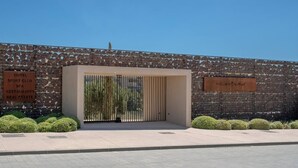 Fachada moderna de un edificio con un portón metálico, un muro de piedra y un cartel que enumera servicios como hotel, club deportivo, spa, restaurantes y bienes raíces.