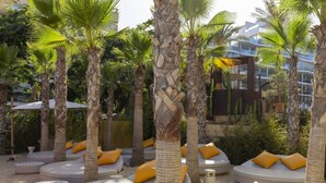 Zonas de estar al aire libre con cojines blancos redondos y almohadones amarillos, rodeadas de palmeras y suelo de arena.