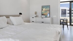 Un dormitorio moderno con una cama grande, un tocador blanco, un escritorio con una silla y un cuadro de un paisaje urbano en la pared.