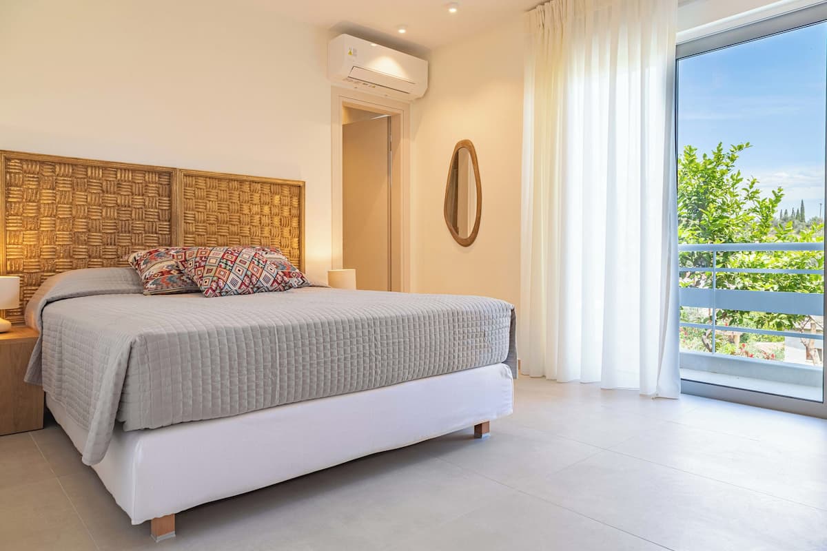 Une chambre moderne avec un grand lit, une tête de lit en bois, une table de chevet, un miroir et un balcon donnant sur un paysage verdoyant.
