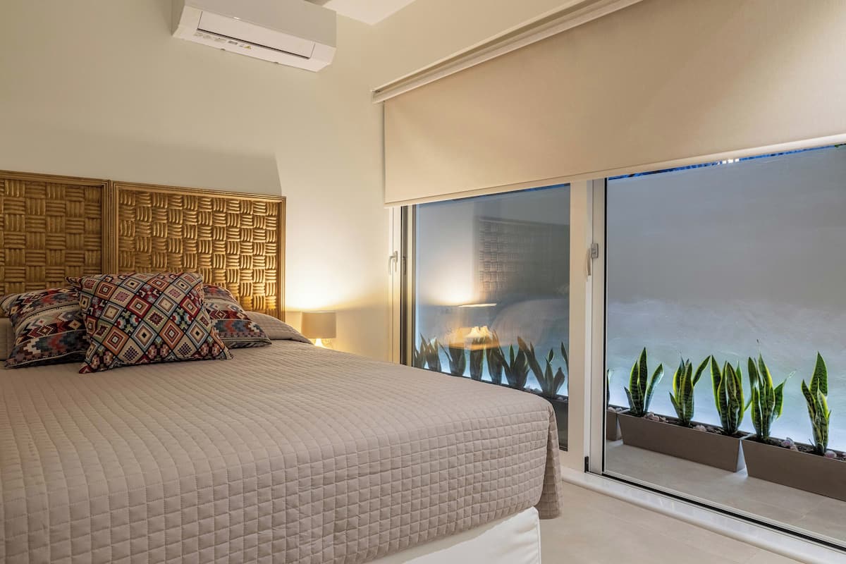 Une chambre avec un lit, des coussins à motifs, un mur orné d’un motif géométrique, une table de chevet avec une lampe et un balcon agrémenté de plantes en pot.