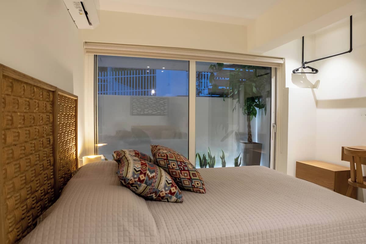 Une chambre avec un grand lit, des coussins à motifs, une tête de lit en bois, une porte-fenêtre coulissante, une table de chevet et un luminaire suspendu.