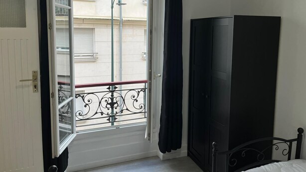 Ein Zimmer mit großem Fenster, einem Balkon mit Metallgeländer, einem schwarzen Kleiderschrank und einem Bett mit Metallgestell.