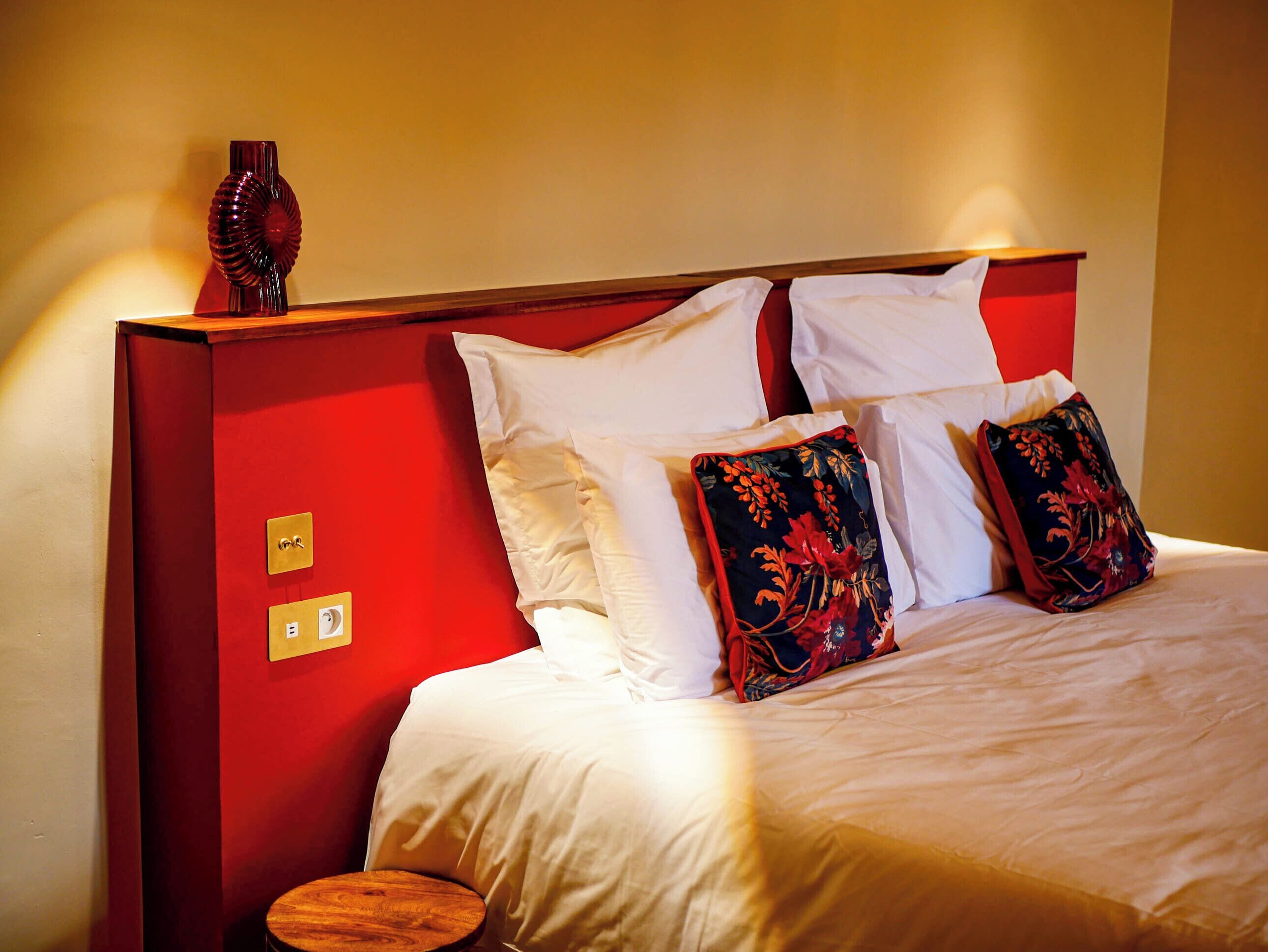 A neatly made bed with white linens and decorative pillows, a wooden bedside table, and a red headboard.