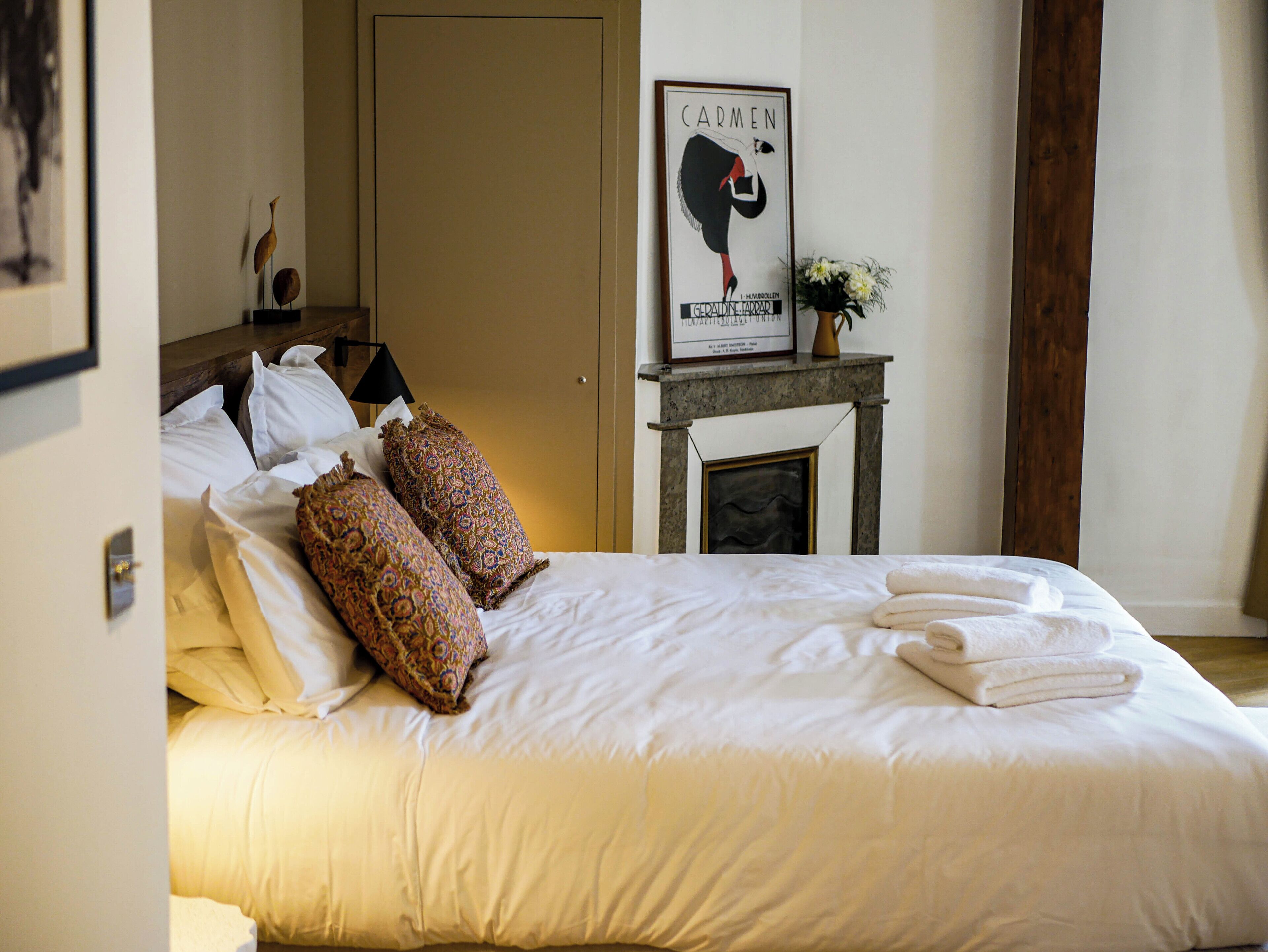 Een net opgemaakt bed met witte lakens en decoratieve kussens, een ingelijste poster aan de muur, en een tafeltje met een bloevas.