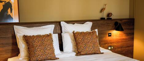 Una cama bien tendida con sábanas blancas y dos almohadas decorativas, una lámpara de pared y un cartel en la pared.