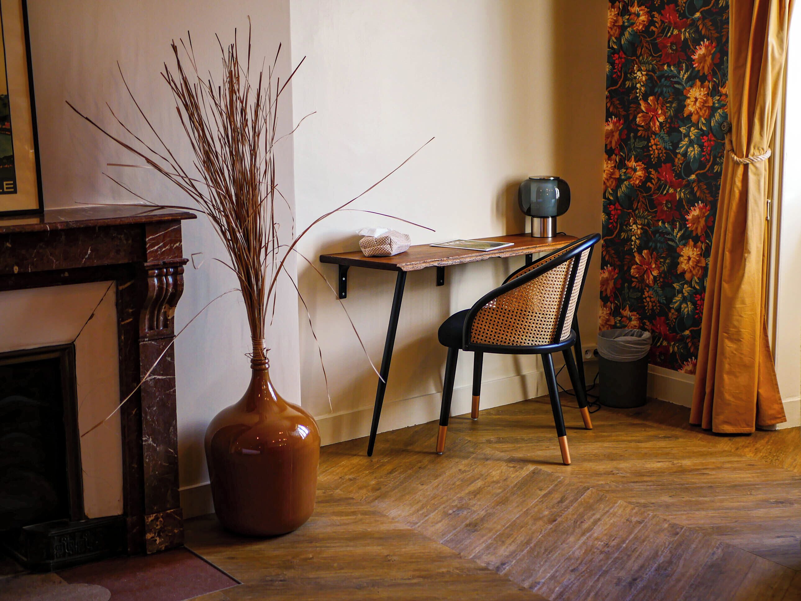 A room with a wooden floor, a chair, a table, a vase with branches, a fireplace, and a curtain with a floral pattern.