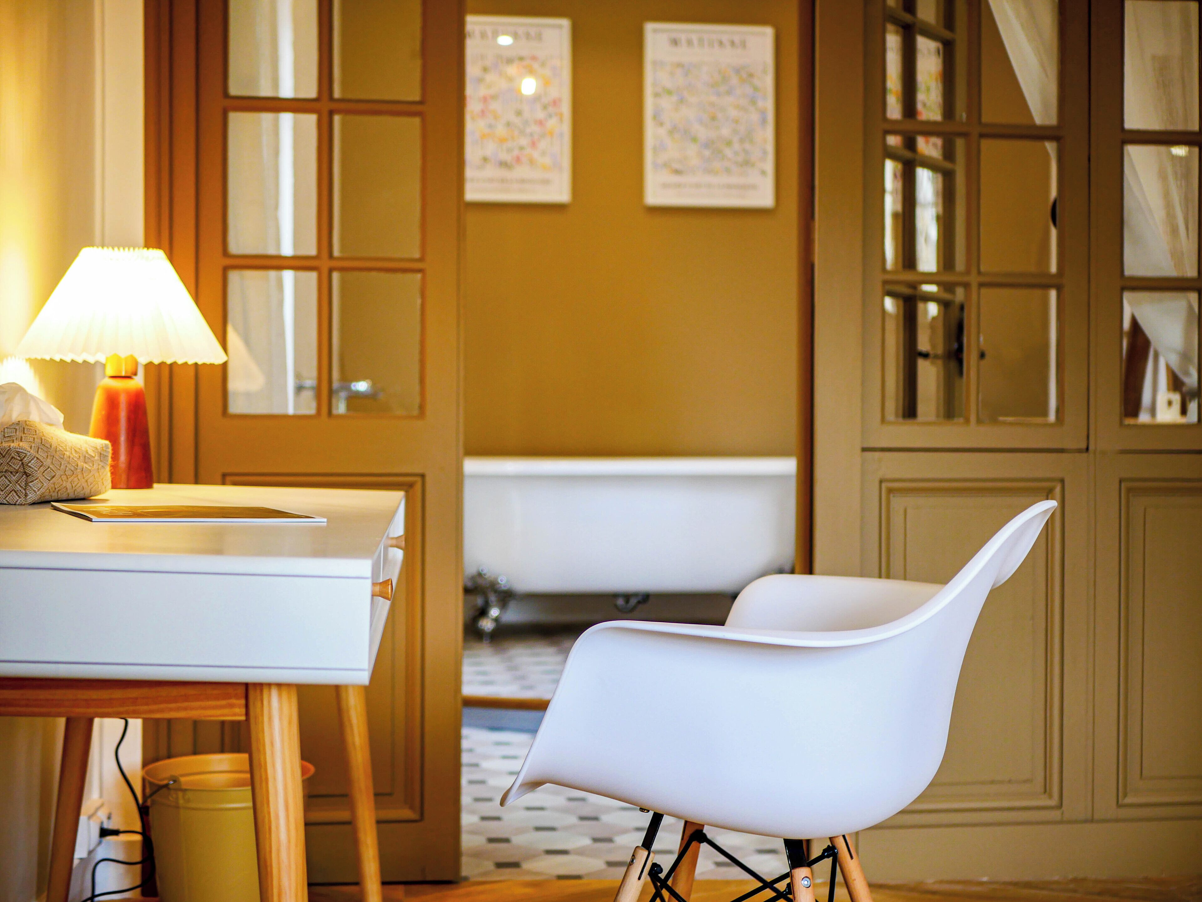 A bathroom with a bathtub, a white chair, a table with a lamp, and a tissue box.