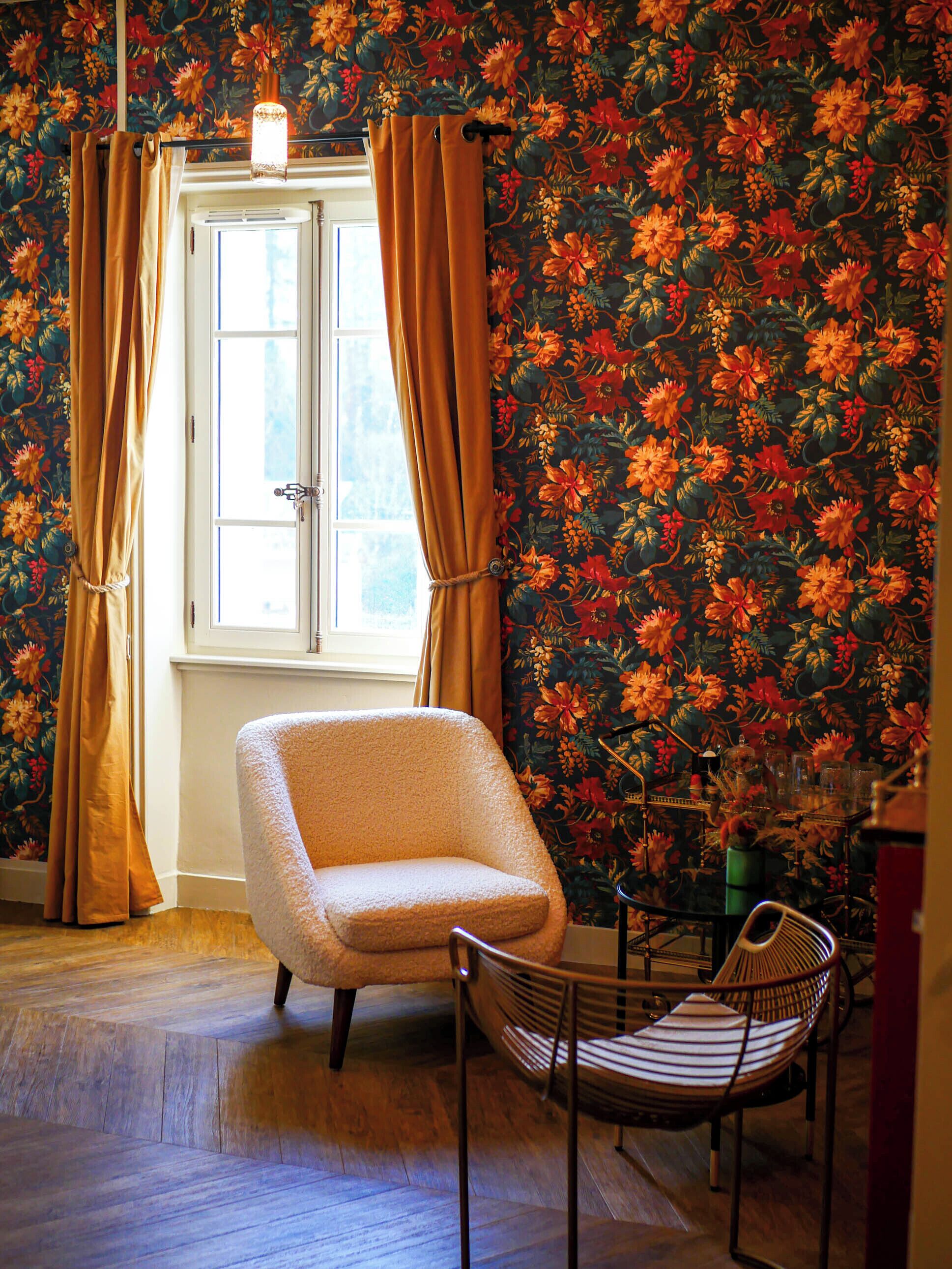 A room with a floral wallpaper, a window with curtains, a chair, and a table with various items.