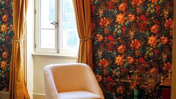A room with a floral wallpaper, a window with curtains, a chair, and a table with various items.