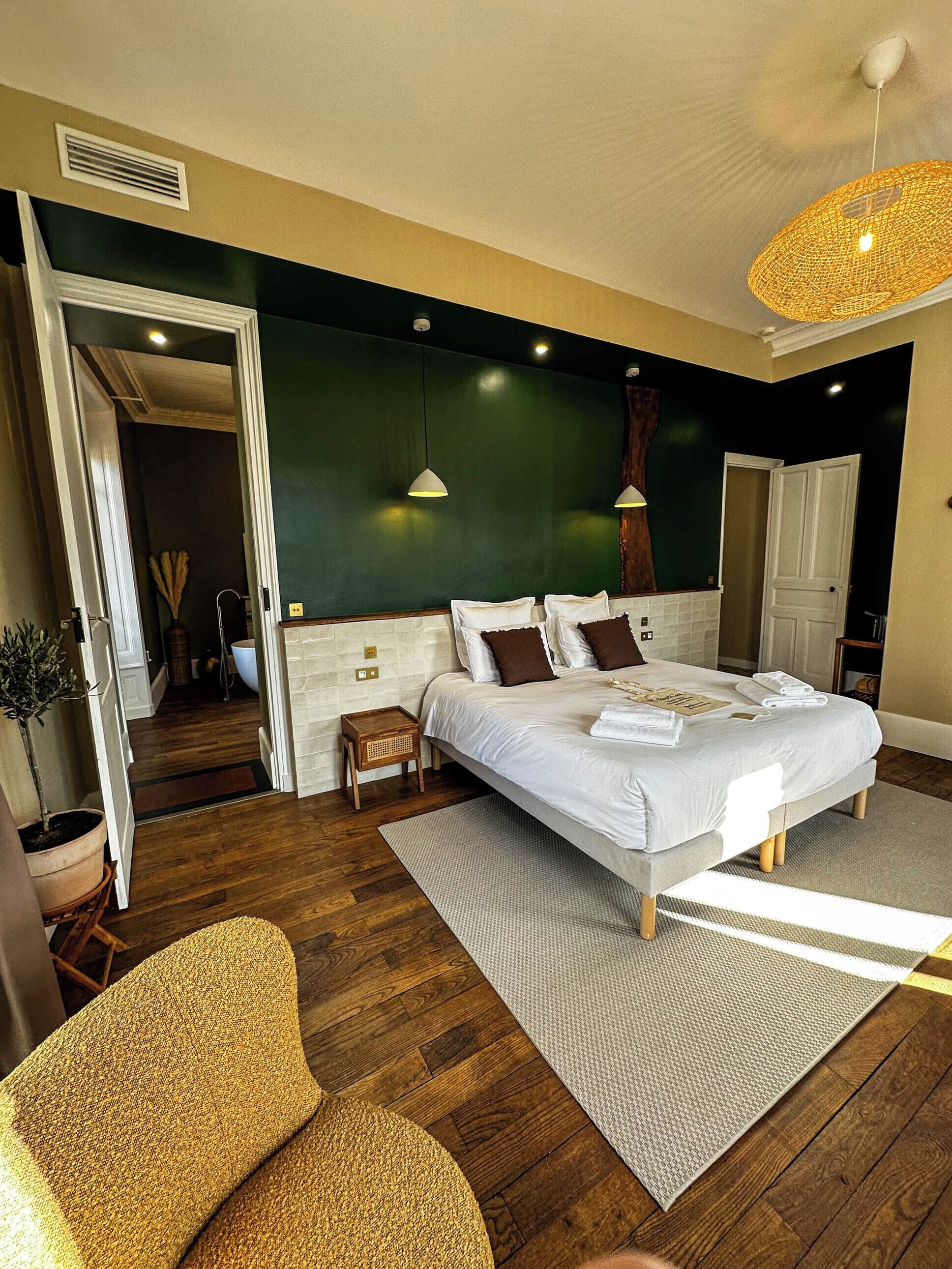 A modern bedroom with a large bed, a small side table, a potted plant, and a view of the bathroom.