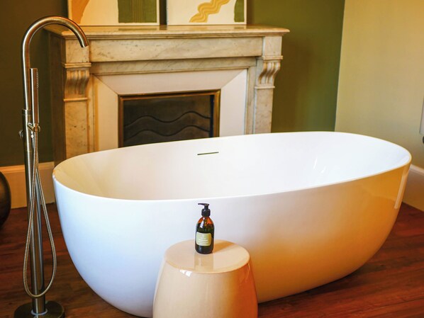 A large oval bathtub with a handheld showerhead, a bottle of shampoo, and a marble pedestal.