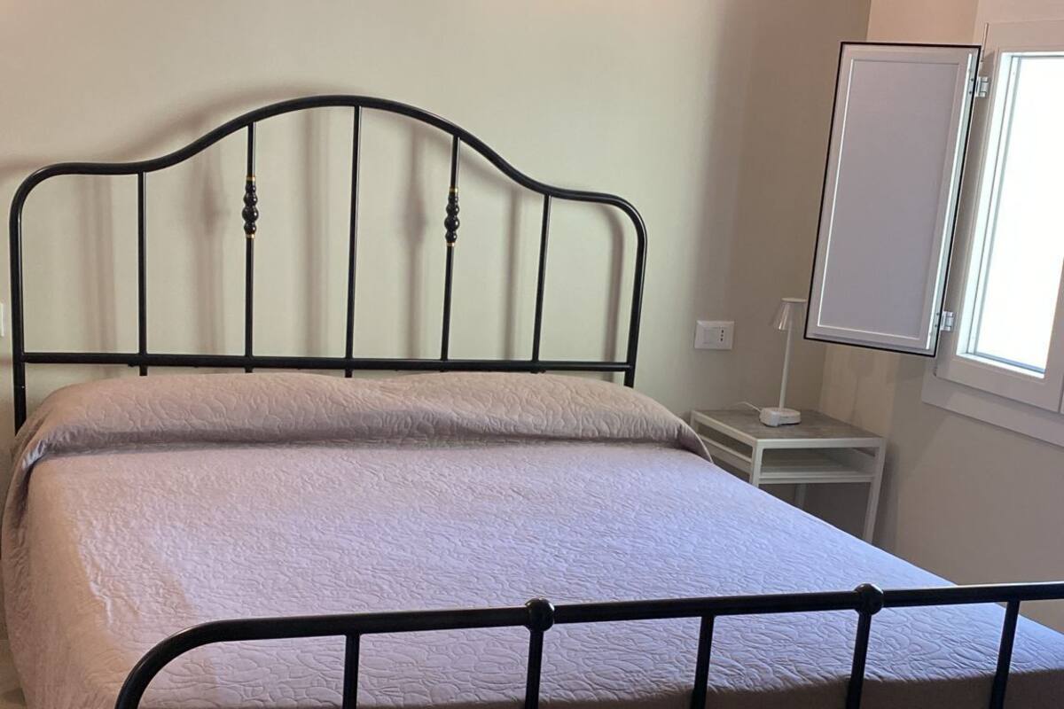 A single bed with a metal frame and a headboard, a bedside table, a wall-mounted light fixture, and a window with blinds.