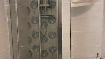Un box de ducha con paredes de vidrio, un cabezal de ducha y un accesorio de ducha de mano, un inodoro con una toalla blanca encima y un rollo de papel higiénico en la pared.
