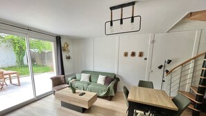 Ett modernt vardagsrum med en grön soffa, ett träbord och ett matbord med stolar. Det finns en trappa med ett träräcke och en skjutdörr i glas som leder ut till ett utomhusområde.