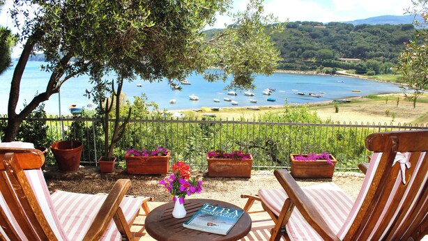 Une terrasse avec deux chaises en bois, une petite table sur laquelle se trouvent un livre et un vase de fleurs, et une vue sur des bateaux sur un lac.