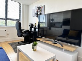 Una sala de estar moderna con un televisor de pantalla plana grande, una silla de oficina negra, una mesa de centro blanca con una planta y una cocina al fondo.