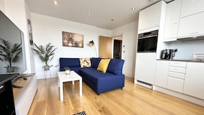 A modern living room with a blue sofa, a white coffee table, and a kitchen area with white cabinets and a black coffee maker. - Condo Apartment With Workspace (Haywards Heath)