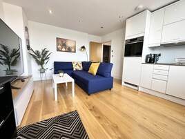 Una sala de estar moderna con un sofá azul, una mesa de centro blanca y una cocina con armarios blancos y una cafetera negra.